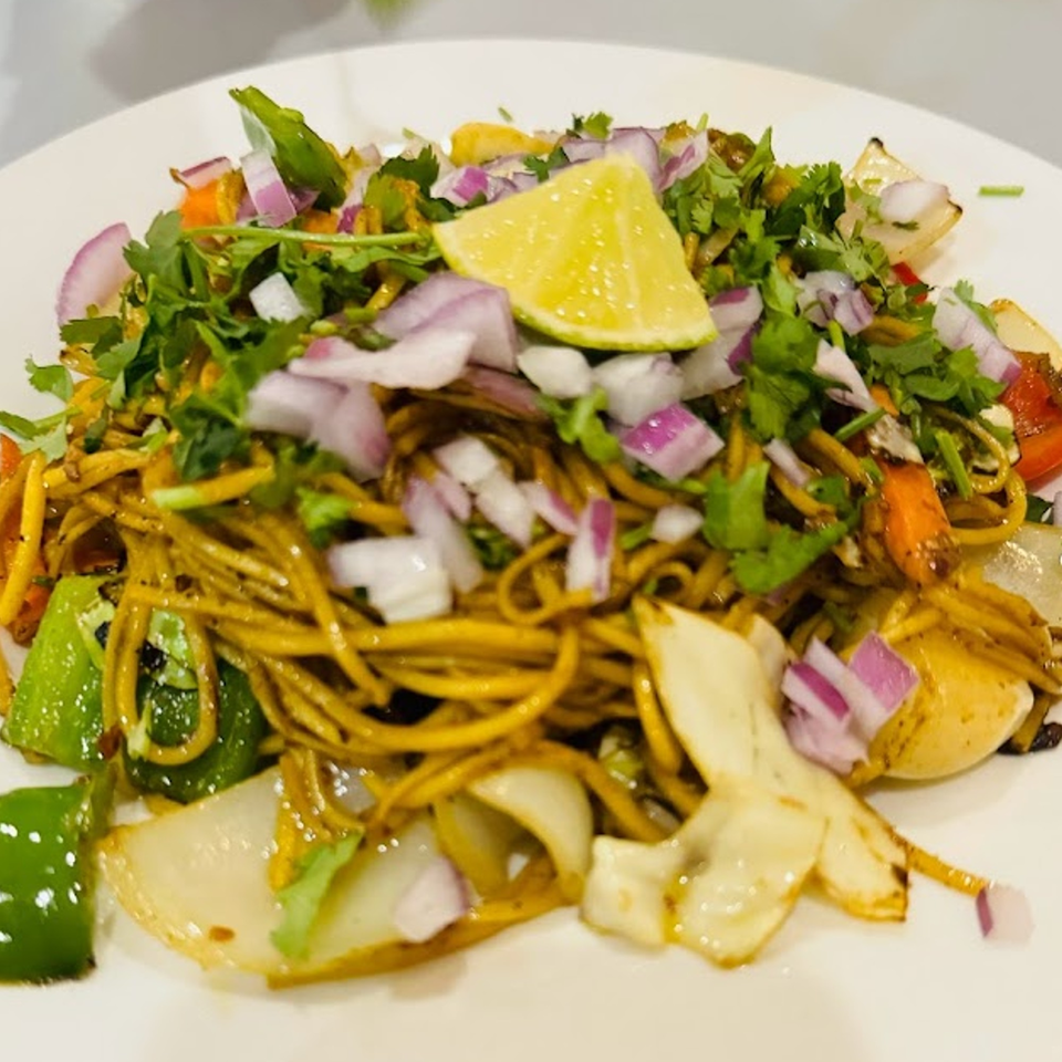 Nepalese chowmein noodles with vegetables, red onions, cilantro, and a lime wedge on a white plate.